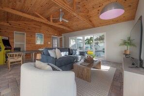 A modern living room with a wooden ceiling, a grey sofa, a white armchair, a wooden coffee table, and a large window with a view of the outdoors. - 4BR Family Oasis - Heated Pool - Walk to Beach (St. Pete Beach)