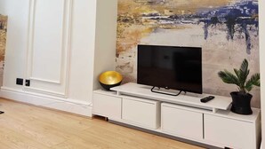 Un salón con un televisor de pantalla plana sobre un mueble blanco, un cuadro abstracto grande en la pared y una planta en maceta.
