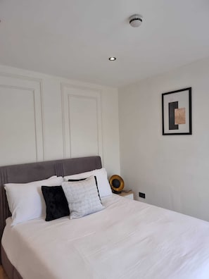 Ein ordentlich bezogenes Bett mit weißer Bettwäsche und einem grauen Kopfteil, ein gerahmtes Bild an der Wand und eine Deckenleuchte.