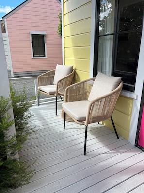 Two wicker chairs with cushions on a wooden deck, a pink building, and a yellow building in the background. - Pinot Villas (Martinborough)