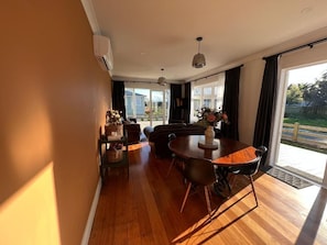 A modern living room with a wooden floor, a dining table with chairs, a sofa, and a large window with curtains.