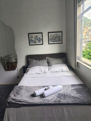 Uma cama bem arrumada com almofadas, uma mesa de cabeceira com abajur e duas fotografias emolduradas na parede.