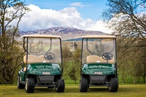 Dos carritos de golf con la marca 'Cally Palace Hotel & Golf Course' estacionados en un campo cubierto de césped, con árboles y montañas al fondo.