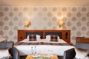 Habitación de hotel con una cama grande, dos lámparas de noche, un cabecero de madera, una mesita de noche, una mesa con una revista y una silla.
