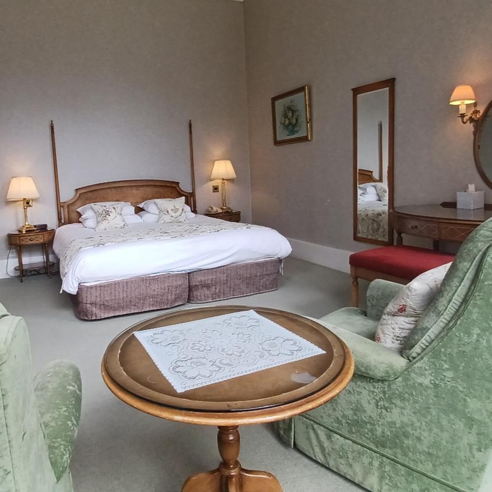 Quarto de hotel com uma cama grande, luminárias laterais, uma mesa redonda, uma poltrona verde e um espelho.