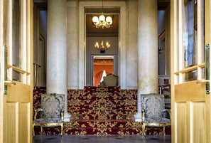 Una imponente escalera con alfombra decorativa y columnas, que conduce a un salón con candelabro.