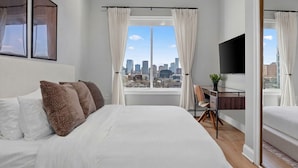 Un dormitorio con cama, escritorio, silla, televisor y vista a la ciudad.