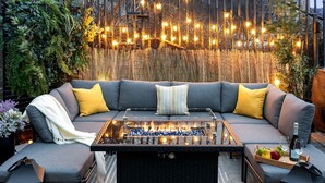 Un espacio al aire libre acogedor con sofá, cojines, una mesa de vidrio y un hogar para fuego.