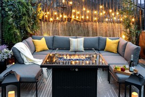 A cozy outdoor seating area with a sofa, cushions, a glass-top table, and a fire feature. - NYC View Patio, Fire Pit, Sleeps 8 (North Bergen)