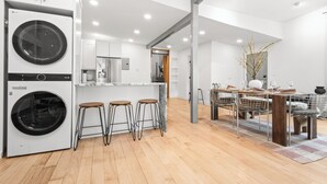 Una cocina moderna con lavadora y secadora integradas, taburetes de bar de madera y un comedor con una silla y una mesa de metal.