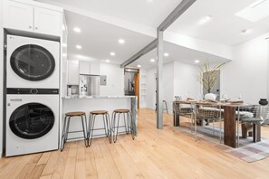 A modern kitchen with a built-in washer and dryer, wooden bar stools, and a dining area with a metal chair and table. - NYC View Patio, Fire Pit, Sleeps 8 (North Bergen)