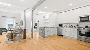 Una cocina moderna con armarios blancos, una isla de mármol, electrodomésticos de acero inoxidable y un comedor con una mesa y sillas de madera.