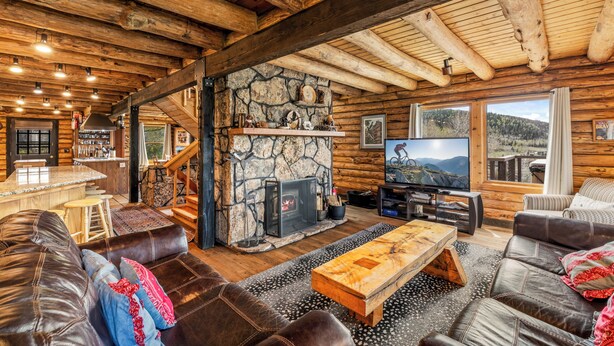 A log cabin interior with a stone fireplace, leather furniture, a television, and a scenic view.