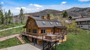 A log cabin with a deck, surrounded by a grassy area and trees, with a mountain in the background.