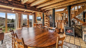 A wooden interior with a large round table, chairs, a fireplace, and large windows offering a view.