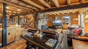 A log cabin interior with a bar area, leather seating, a stone fireplace, and a mountain view.