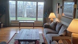 A living room with a large window, a sofa, a coffee table, and two lamps. - Creekside Retreat at Notre Dame (South Bend)