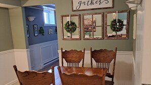 A dining area with a wooden table and chairs, a wall-mounted sign that says 'gathers,' and framed mirrors with greenery. - Creekside Retreat at Notre Dame (South Bend)
