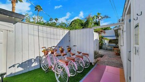 Een omheinde achtertuin met een rij roze fietsen, een klein terras en een wit gebouw met een deur.