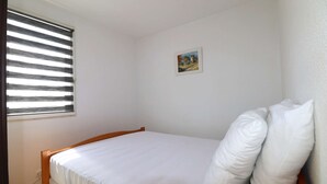 A single bed with white bedding, a small window with horizontal blinds, and a framed picture on the wall. - 2-room apartment for 4 people, 5 minutes from the beach (LE GRAU DU ROI)