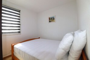 Ein Einzelbett mit weißer Bettwäsche, ein kleines Fenster mit Rollläden und ein gerahmtes Bild an der Wand.