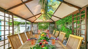 A covered outdoor dining area with a long table, chairs, and hanging plants. - Spirit of Sofia 12bd (Palm Springs)