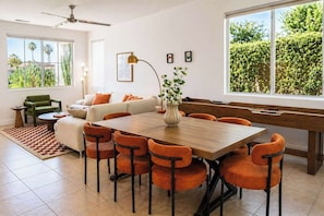 A dining area with a wooden table and orange chairs, a sofa with orange cushions, a green chair, a floor lamp, and a large window with a view of greenery.