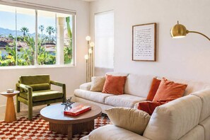 A living room with a sofa, armchair, coffee table, and a view of greenery outside.