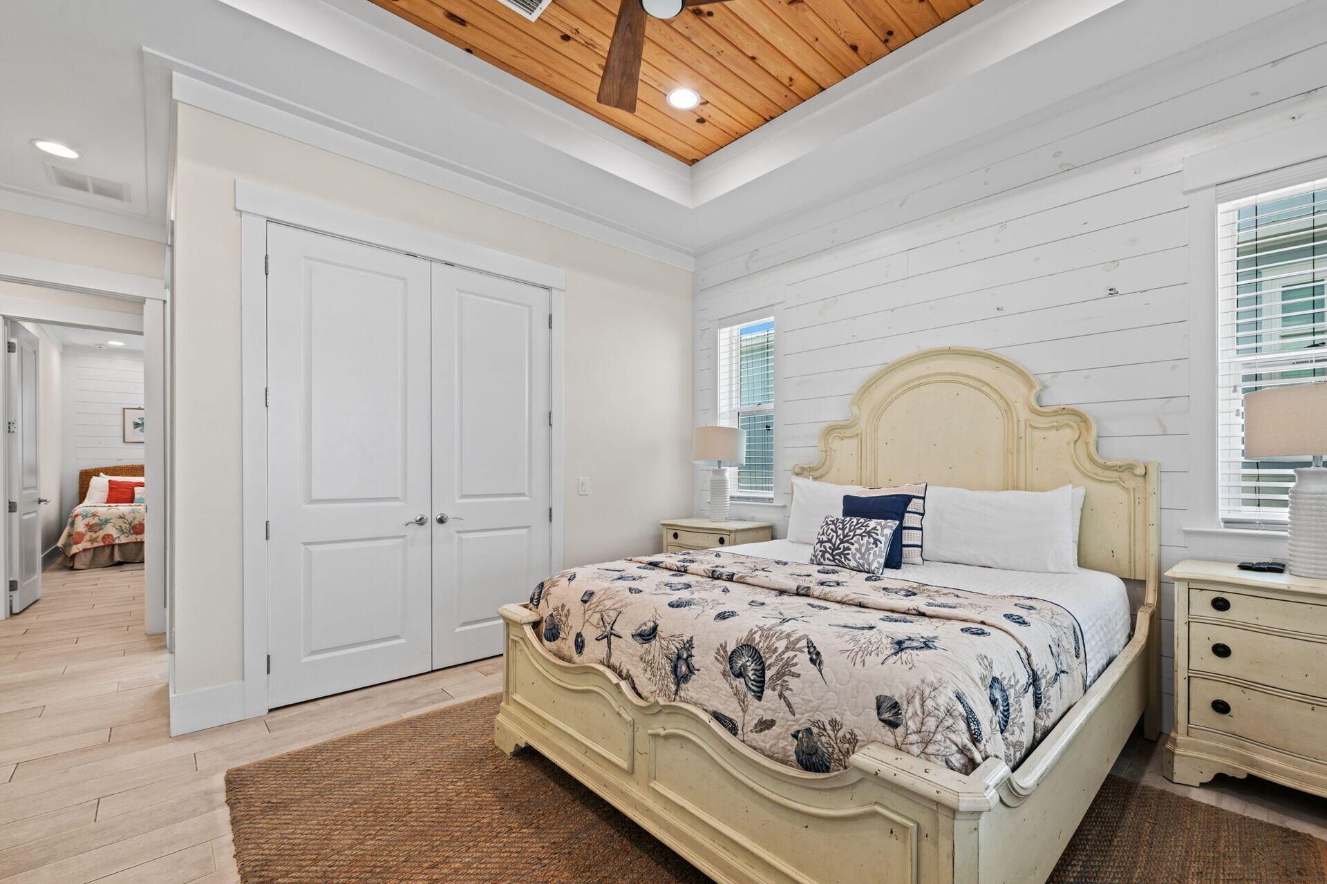 Une chambre avec un plafond en bois, un grand lit recouvert d’une couvre-lit à motifs floraux, des murs blancs et une fenêtre avec des stores.