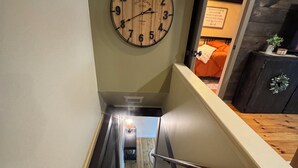 Un escalier avec une rampe en bois, une horloge fixée au mur et un aperçu d’une pièce où l’on aperçoit un lit et une plante.