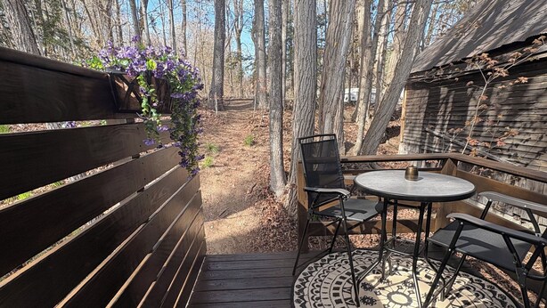 Une terrasse en bois avec un banc et une table entourée de chaises, le tout bordé d’arbres.