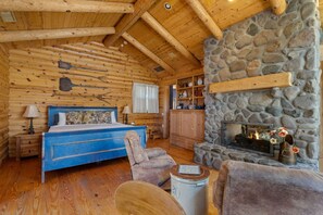 Une chambre située dans un chalet en rondins, avec une cheminée en pierre, des meubles en bois et un lit agrémenté d’une literie à motifs floraux.