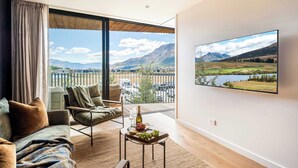 En moderne stue med et stort vindue, en sofa, en lænestol, et lille bord med en flaske og druer, og et fjernsyn der viser et landskab.