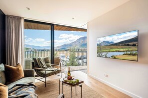 Ein modernes Wohnzimmer mit einem großen Fenster, einem Sofa, einem Sessel, einem kleinen Tisch mit einer Flasche und Trauben sowie einem Fernseher, der eine Landschaft zeigt.