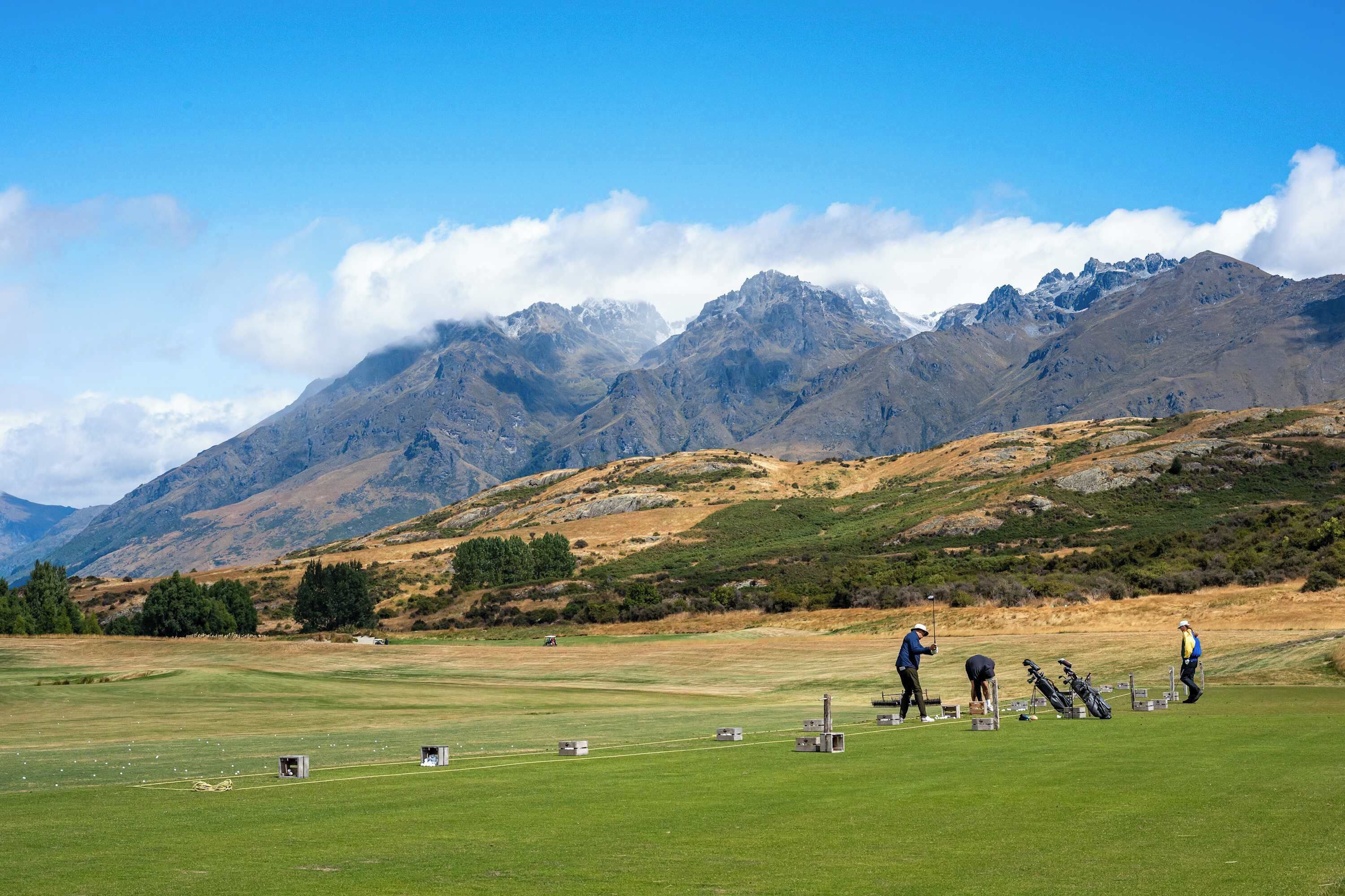 A golf course with players and equipment, surrounded by mountains and clear skies.