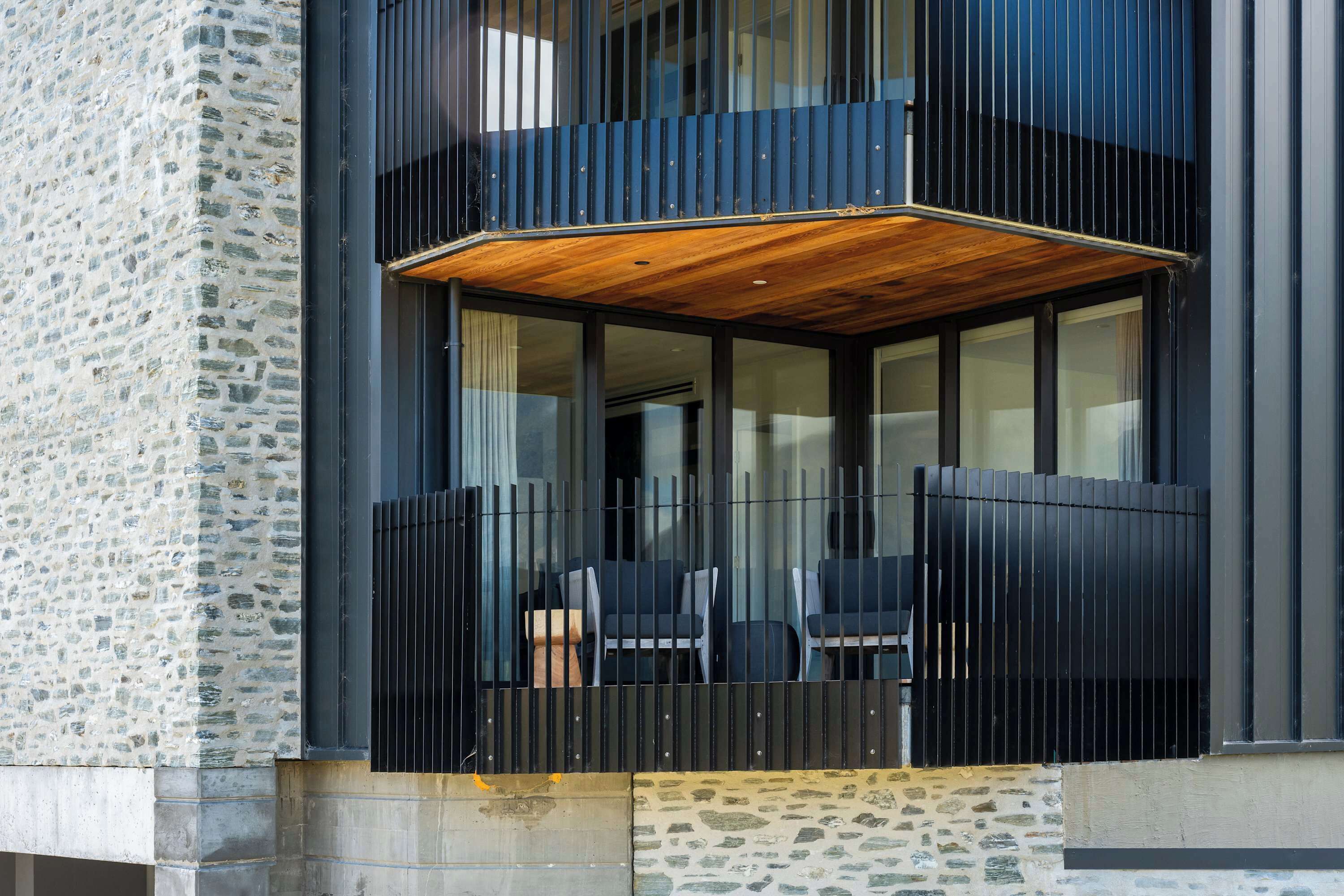 A modern building with a stone facade, a balcony with wooden flooring, and black-framed glass windows.