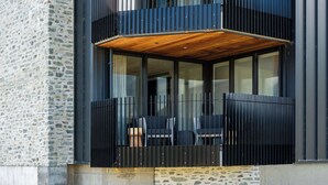 Un bâtiment moderne avec une façade en pierre, un balcon doté d’un plancher en bois et des fenêtres à vitres encadrées de noir.
