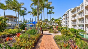 A resort with a pool, palm trees, and a brick pathway. - Sun & Surf Pad Resort Living, Alexandra Headland (Alexandra Headland)