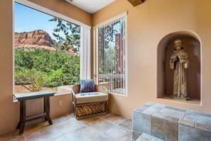 A room with a view of red rock formations, a stone statue, a cushioned chair, and a small table. - Purple Lizard- Sedona Villas (Sedona)