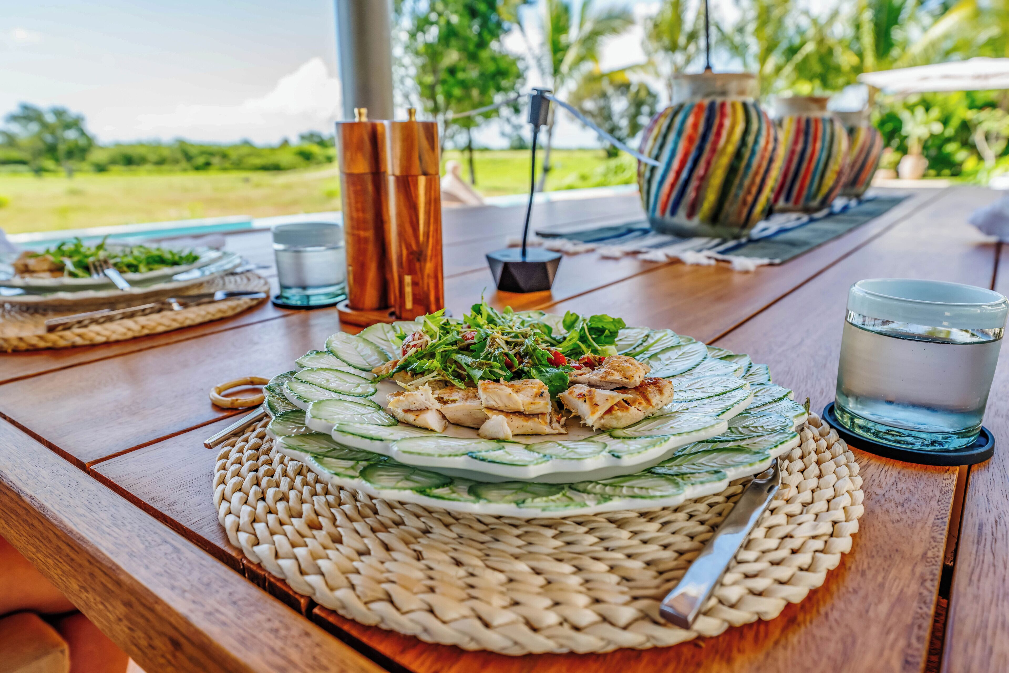 Une table en bois, avec une assiette de nourriture, un verre d’eau et un set de table en fibres tressées.