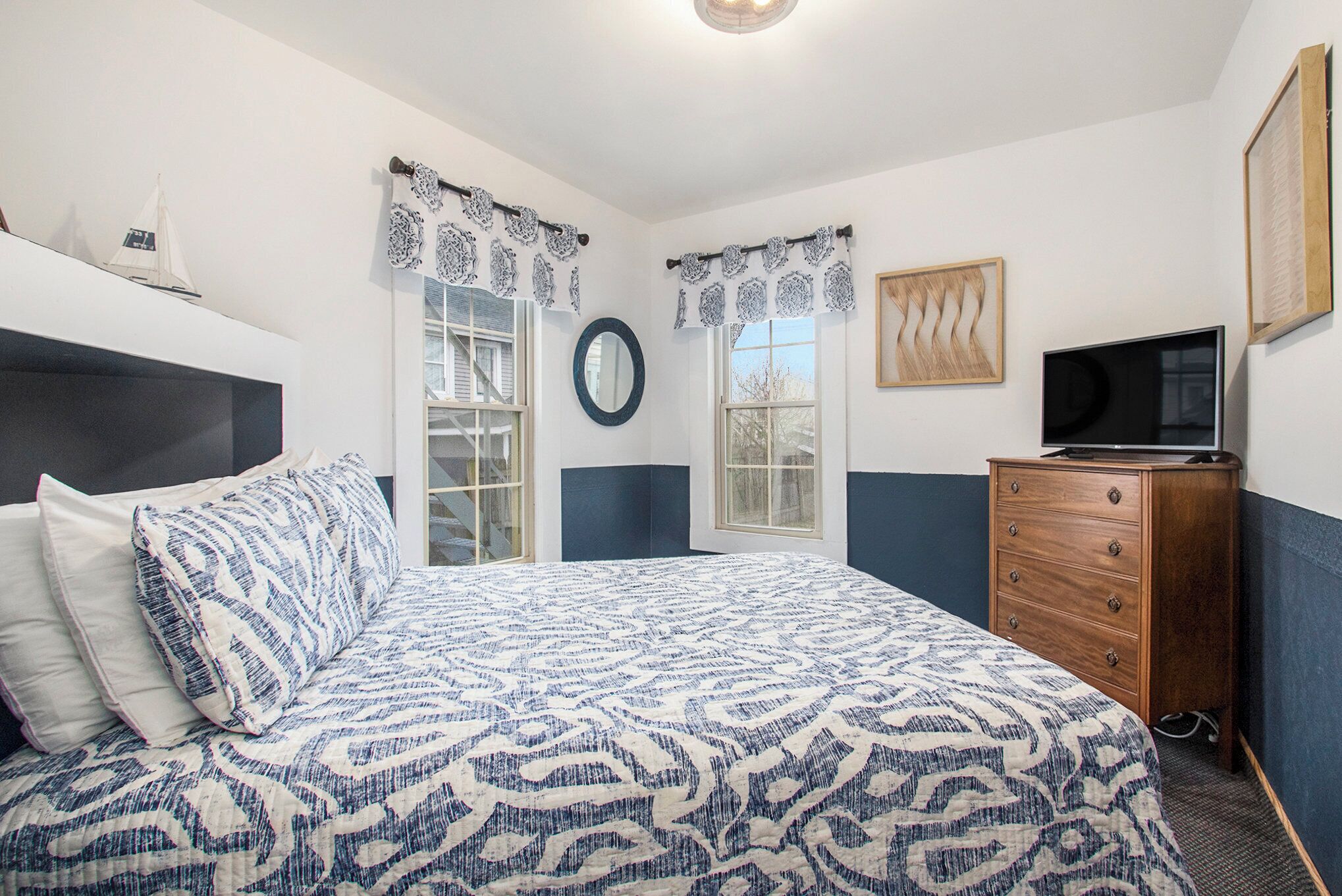 A bedroom with a bed, dresser, TV, windows, and curtains.
