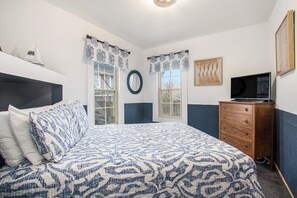 Ein Schlafzimmer mit Bett, Kommode, Fernseher, Fenstern und Vorhängen.
