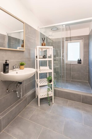 A bathroom with a white sink, a mirror, a glass shower enclosure, and a shelf with toiletries. - Villa Rheinsteig Sauna-whirlpool-kino (Bendorf)