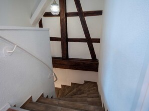 Eine Treppe mit Holzboden und weißen Wänden führt zu einem Raum mit schrägem Dach und einer Pendelleuchte.