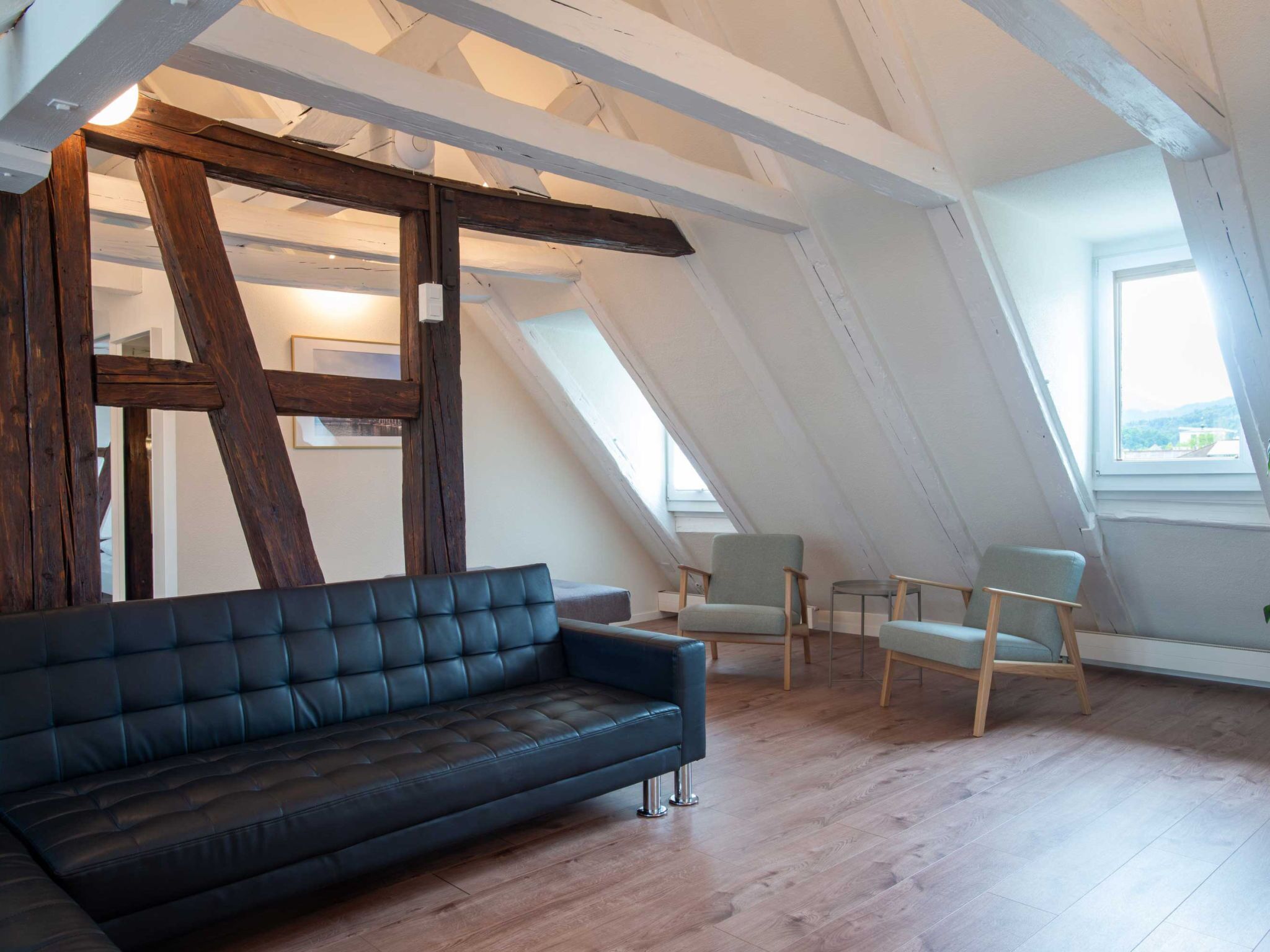 Ein Loft-Zimmer mit einem dunkelbraunen Ledersofa, zwei hellen Sesseln und einer Holzbalkendecke.