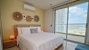 Ein Hotelzimmer mit Bett, Nachttischlampen, einer fest installierten Klimaanlage und einem großen Fenster mit Blick auf ein Hochhaus und den Strand.