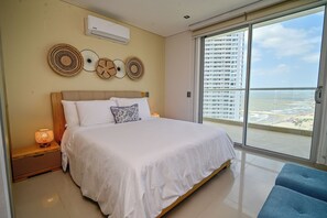 A hotel room with a bed, bedside lamps, a wall-mounted air conditioner, and a large window with a view of a high-rise building and beach. - Luxury apartment in cartagena| design +comfort (Cartagena de Indias)