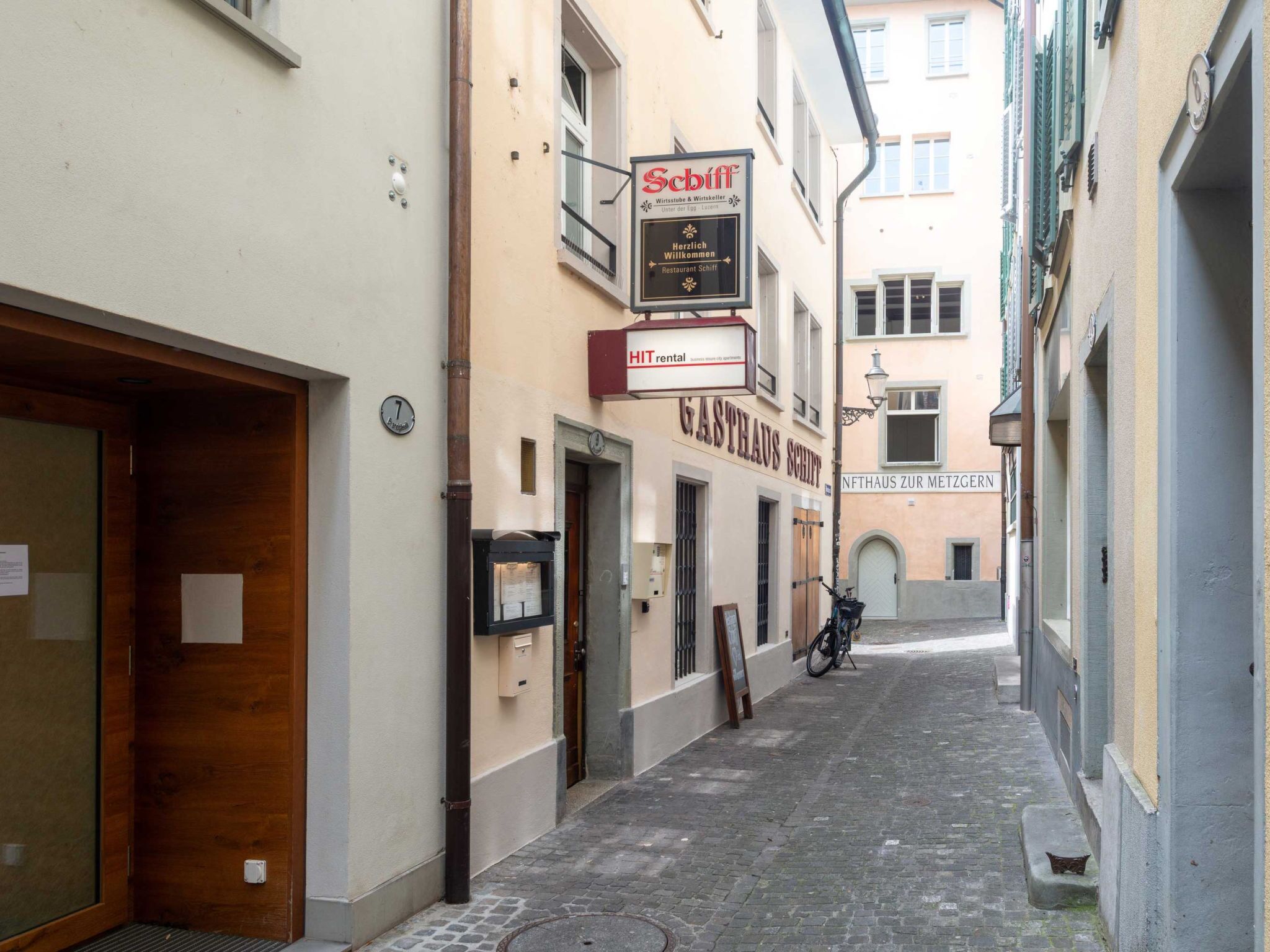 Una calle estrecha flanqueada por edificios, incluyendo un restaurante llamado 'Schiff' y un letrero de alquiler de autos 'HIT'.