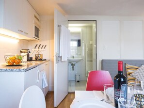 A modern kitchen with white cabinets, a countertop with fruit bowl, and a dining area with a red chair and table set for two. - Apartment  by Interhome (Zoug)