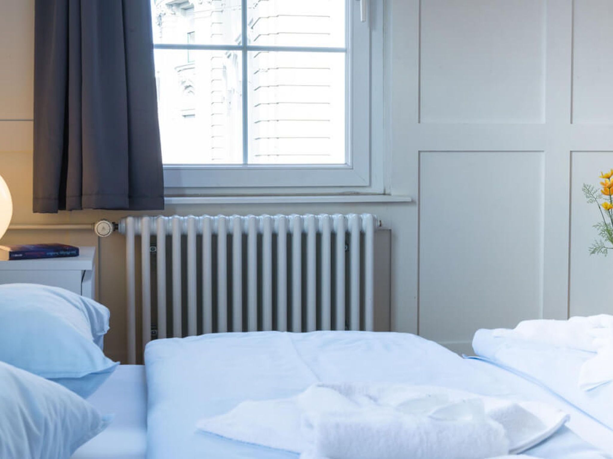 Une chambre d’hôtel avec un lit, une table de chevet avec une lampe, une fenêtre avec des rideaux, un radiateur et un mur orné d’un tableau.